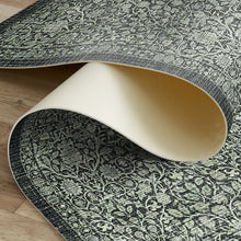 Load image into Gallery viewer, A close-up of a rolled area rug showing its dark gray and off-white floral damask pattern and cream-colored underside, laid over a light wood floor.