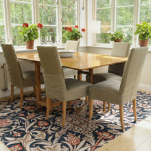 Load image into Gallery viewer, Dining room with wooden table and chairs near large windows with plants.