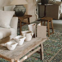 Load image into Gallery viewer, Three small handmade off-white ceramic vessels with integrated handles sit on a rustic wooden coffee table in a cozy living room setting with a patterned rug, a white sofa, and a side table with a lamp.