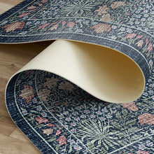 Load image into Gallery viewer, A close-up of a rolled area vinyl rug showing its navy blue floral pattern with pink and beige accents, alongside its cream-colored underside, placed on a light wood floor.