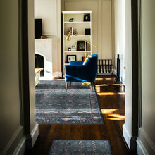 Load image into Gallery viewer, A blue patterned area rug is shown in a real home interior, viewed through a doorway. The rug covers part of a dark wood floor in a living room with a blue armchair, white bookshelf, fireplace, and sunlight streaming in.