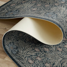 Load image into Gallery viewer, A close-up of a patterned area rug being rolled up, showing both the top and bottom sides. The top side features a dark navy blue and gray botanical pattern with muted pink accents, while the underside is a solid cream color.