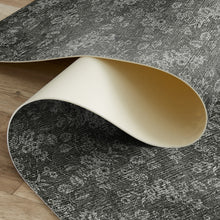 Load image into Gallery viewer, A dark gray and white distressed floral patterned rug partially rolled up to show its cream-colored underside, displayed on a light wood floor.