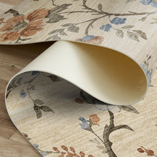 Load image into Gallery viewer, A close-up of a rolled area rug showing its cream-colored underside and a floral pattern on top. The rug features a textured beige background with branches and flowers in muted shades of brown, blue, and gray, displayed on a light wood floor.
