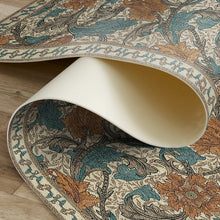 Load image into Gallery viewer, A rolled section of a traditional area rug with an ornate floral and scroll pattern in beige, teal blue, and rust orange, shown on a light wood floor.

