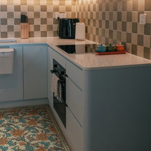 Load image into Gallery viewer, A modern kitchen corner with light blue cabinets, a white countertop, a beige and gray checkered tile backsplash, a built-in black oven, and decorative kitchen accessories including colorful teapots and a paper towel holder.
