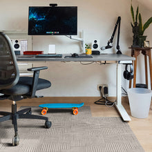 Load image into Gallery viewer, A modern home office setup featuring a gray under-desk monitor arm holding a large desktop monitor, with a black office chair, speakers, desk accessories, a blue skateboard on a gray patterned rug, and a potted plant on a wooden side table.
