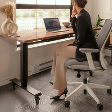 Load image into Gallery viewer, A woman sits in a modern gray and white office chair at a standing wooden desk by a window, with a laptop open on the desk and decorative items nearby.
