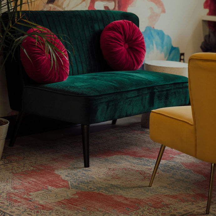 Green velvet bench with pink cushions in a room with a yellow chair and colorful boho rug.