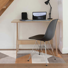 Load image into Gallery viewer, A modern black molded plastic chair with a chrome wire base sits in front of a light wood desk in a home office setting. The desk holds a laptop, notebook, and black desk lamp, set against a white wall with a staircase visible in the upper left.
