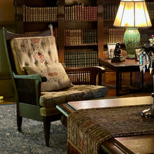 Load image into Gallery viewer, A vintage upholstered armchair with a floral patterned cushion and matching pillow, set in a warm, traditional library with wooden bookshelves, a table lamp, and a patterned rug.
