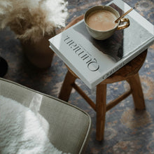 Load image into Gallery viewer, Cup of coffee on a book with a spoon, placed on a wooden stool with a textured rug background.
