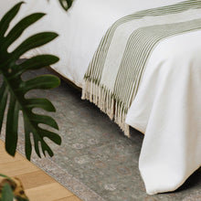 Load image into Gallery viewer, Bed with a green and beige striped blanket on a patterned rug, next to a plant.
