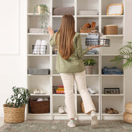Person organizing shelves in a room with various items including plants and baskets. standing on a boho style mat