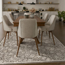 Load image into Gallery viewer, A dining room scene showing a rectangular wooden dining table surrounded by six upholstered chairs with beige textured seats and brown wooden legs, placed on a large floral area rug in a modern kitchen-dining interior.
