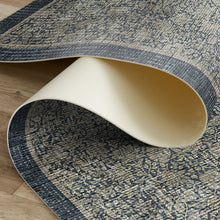 Load image into Gallery viewer, A close-up of a rolled area rug showing its distressed navy blue and beige patterned top side and a cream underside, displayed on a light wood floor.

