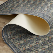 Load image into Gallery viewer, A close-up of a patterned area rug partially rolled up to show both the top woven design and the underside. The rug features a dark blue-gray base with beige, rust, and muted orange floral motifs, laid over a light wood floor.
