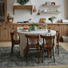 Load image into Gallery viewer, A dining area in a rustic kitchen with a round table covered in a light beige tablecloth, surrounded by four wooden spindle-back chairs. The scene is decorated with a white floral area rug, a green plant, and various kitchen items in the background.
