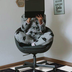 Person wearing a onesie with cat patterns sitting on a black office chair, holding a laptop above their head.