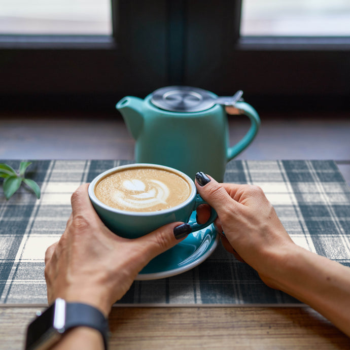 Person holding a blue mug with a cappuccino and a teal teapot on a checkered tablecloth.