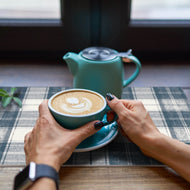 Person holding a blue mug with a cappuccino and a teal teapot on a checkered tablecloth.