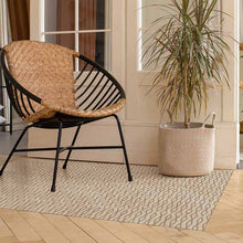 Load image into Gallery viewer, Wicker chair with black metal frame on a patterned vinyl rug next to a potted plant.
