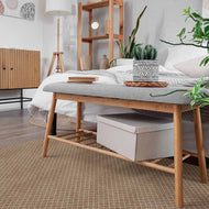Wooden bench with a gray cushion in a bedroom setting with plants and furniture on a jute vinyl mat